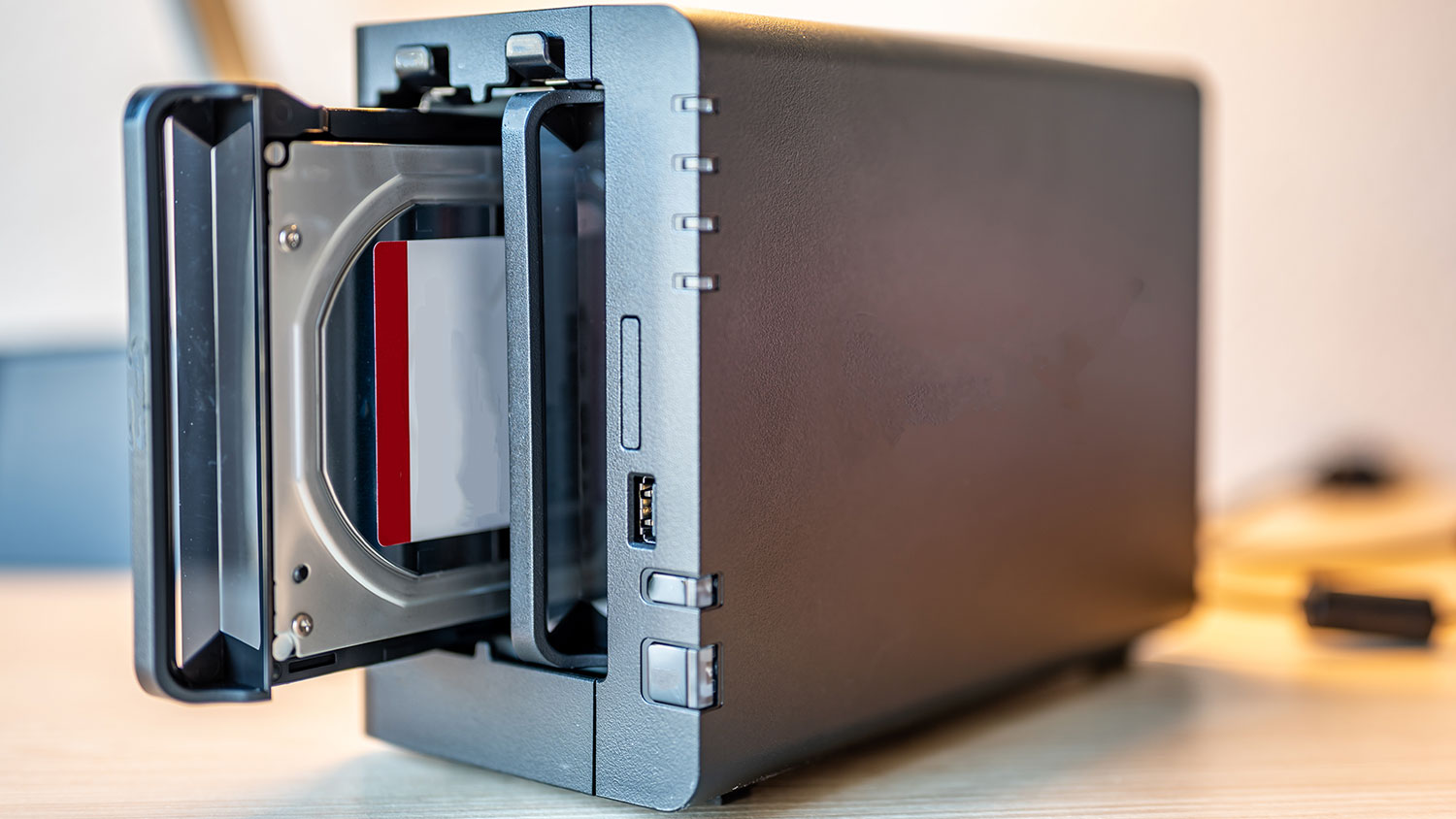 A NAS enclosure sits on a wooden table with one drive tray partially pulled out, revealing an installed hard drive.