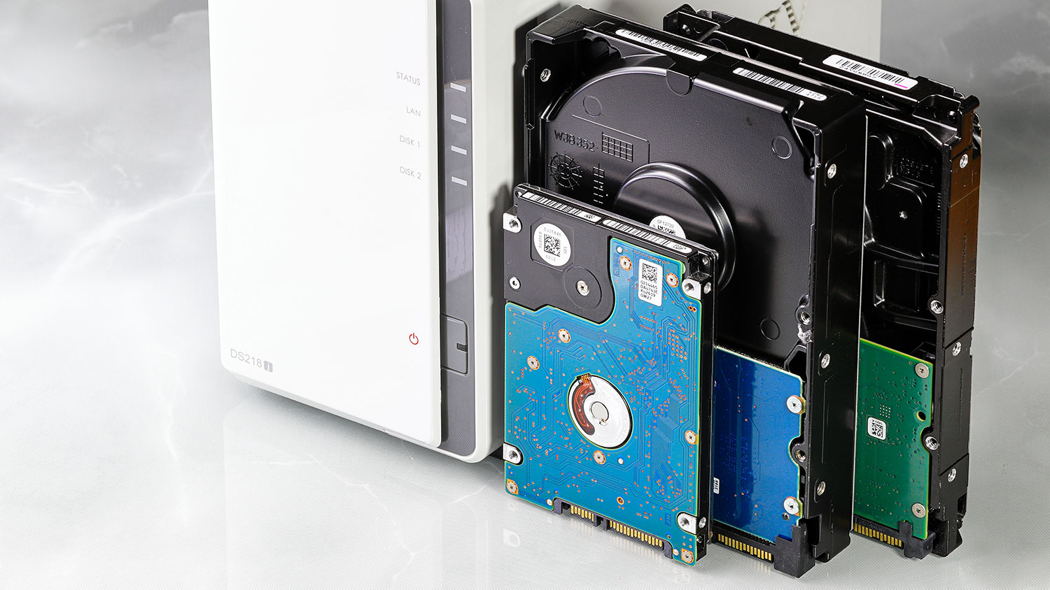 A 2.5-inch hard drive and two 3.5-inch hard drives with exposed circuit boards next to a white NAS storage enclosure on a light surface. A 2.5-inch hard drive and two 3.5-inch hard drives with exposed circuit boards next to a white NAS storage enclosure on a light surface.