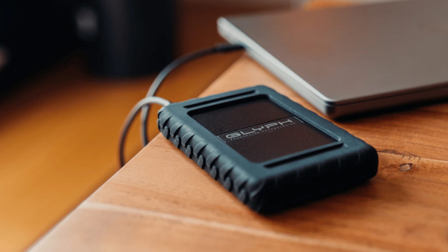 A blue Glyph Rugged external hard drive with a textured bumper that  is connected by a cable to a silver laptop computer on a wooden desk